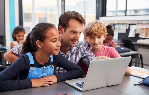 Teacher And Two Young Students Use Laptop Computer In Class